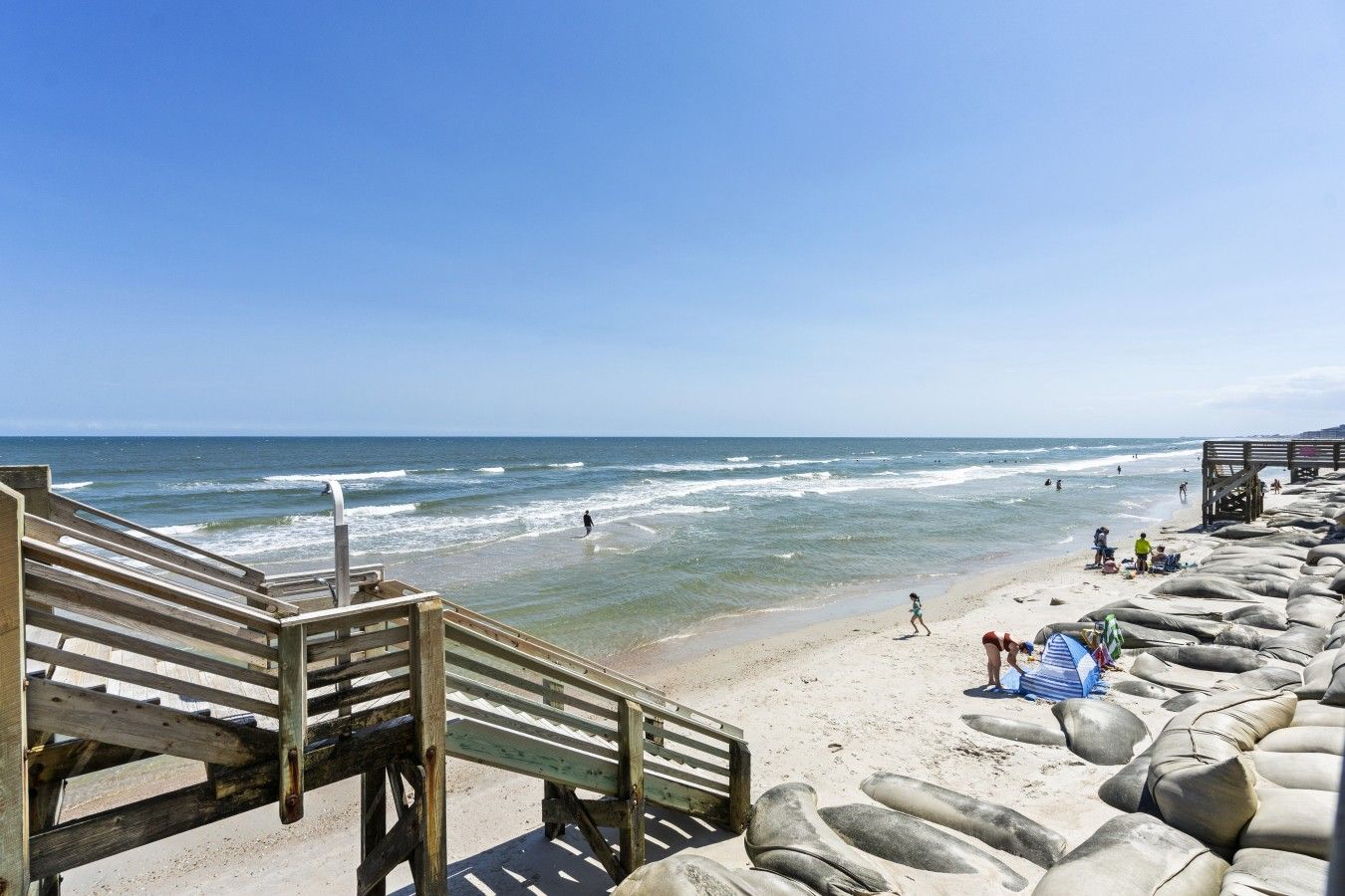 A beach with a wooden staircase leading down to the shore, a sandy beach with rocks, and the ocean in the background.