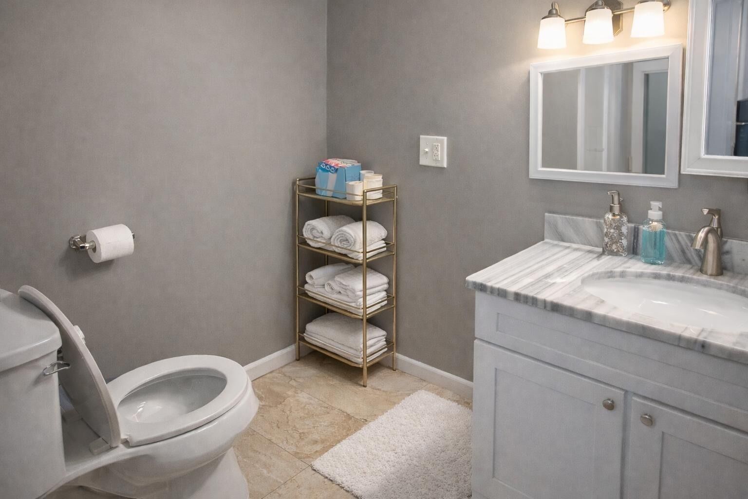 Un baño con inodoro, lavabo con encimera de mármol, toallero con toallas dobladas y espejo sobre el lavabo.