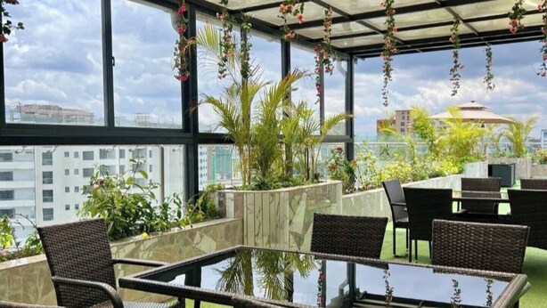 Eine Dachterrasse mit einem Glastisch, umgeben von Korbstühlen, künstlichem Rasen und hängenden Blumenarrangements.