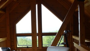 A cozy cabin interior with a wooden ceiling, a large window offering a view of the outdoors, a leather sofa, and a white armchair.