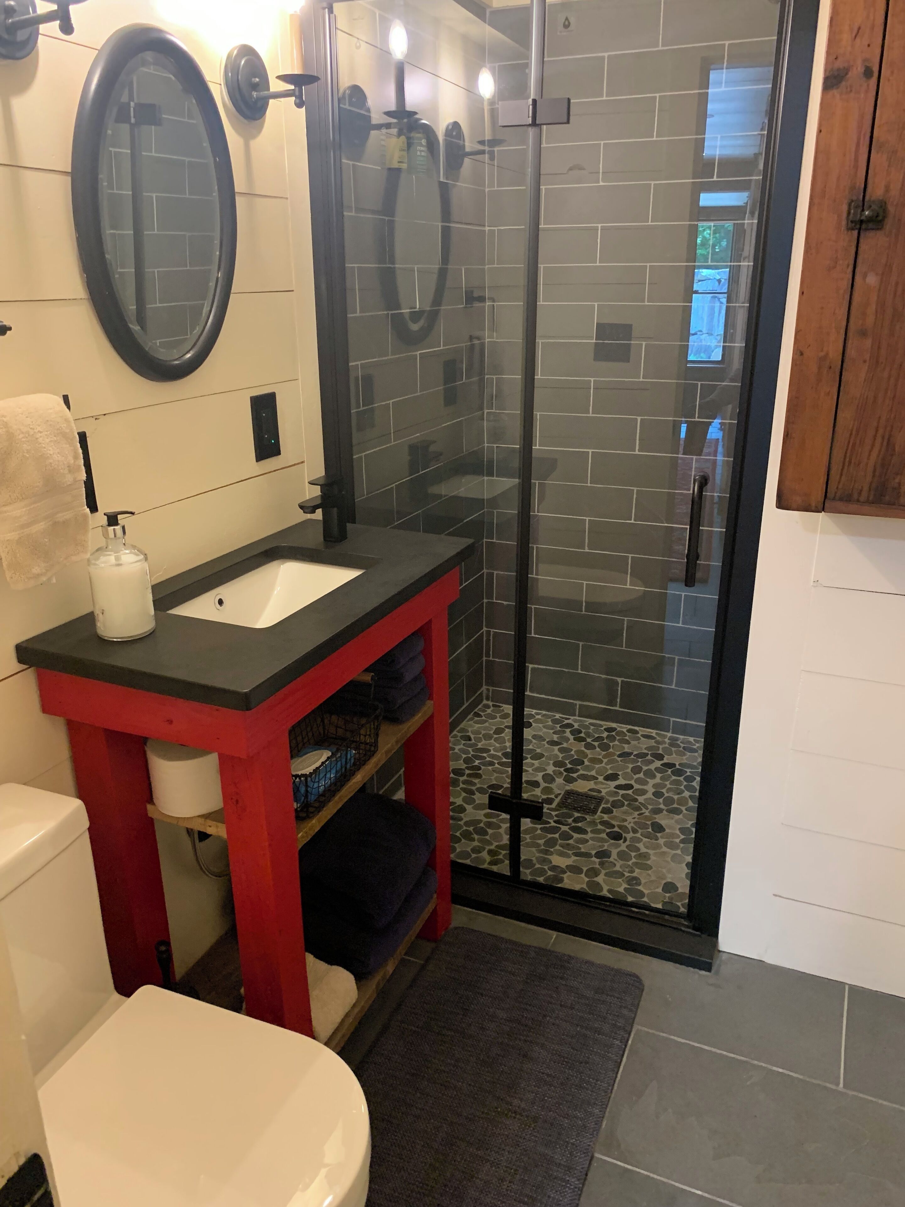 A bathroom with a glass-enclosed shower, a white toilet, a red vanity with a black countertop, and a mirror above the sink.