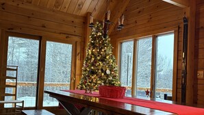 A wooden cabin interior with a dining table, benches, a Christmas tree, and large windows.