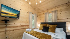 A wooden cabin bedroom with a bed, curtains, a TV, and a window. - Private & Modern Creekside Cabin w/ Hot Tub and Fire Pit - Great Location! (Cherry Log)