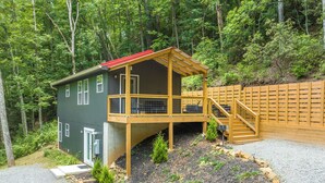 A two-story cabin with a wooden deck and a red roof, surrounded by dense forest. - Private & Modern Creekside Cabin w/ Hot Tub and Fire Pit - Great Location! (Cherry Log)