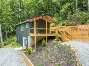 Una cabaña de dos pisos con terraza de madera y techo rojo, rodeada de un bosque denso.