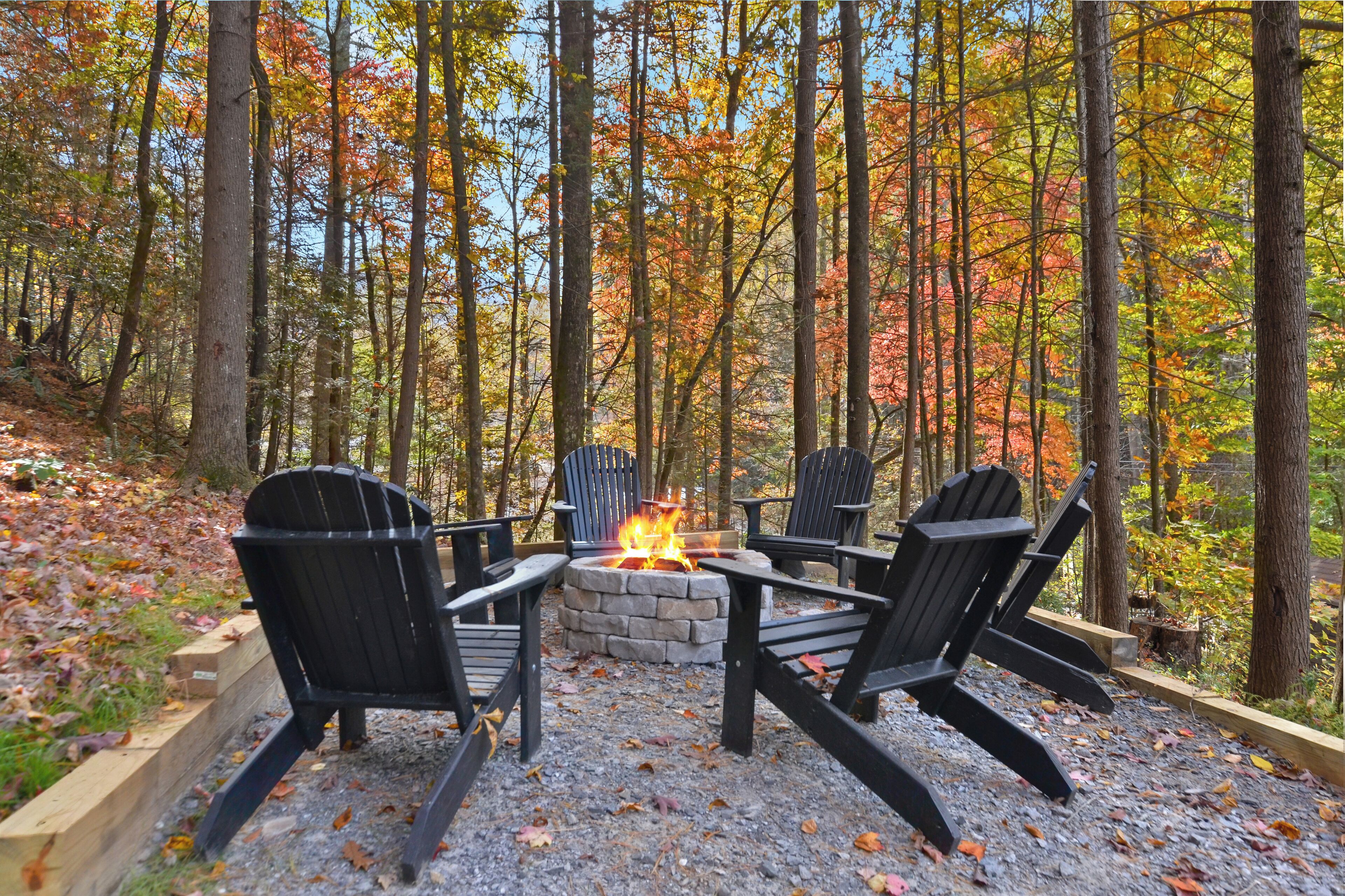 A forest setting with a stone fire pit and four Adirondack chairs arranged around it.