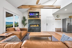 A modern living room with a fireplace, leather sofa, wooden coffee table, and a television mounted above the fireplace. - New from 5 Star Hosts-Panoramic Mtn Views-Private-Peaceful on 5 acres-Convenient (Weaverville)