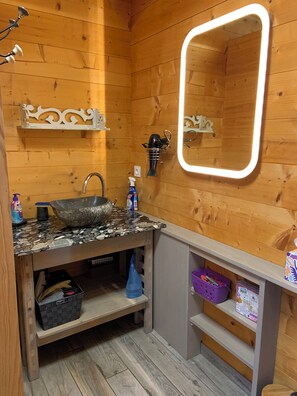 Un interior de madera con lavamanos de encimera de granito, un espejo con luz y estantes con artículos de aseo.
