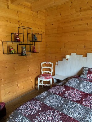Una habitación de madera con una cama, una silla blanca, una mesita y un estante de pared con adornos.