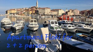 Egy kikötő, ahol a hajók kikötve vannak, a háttérben egy városképpel, és egy felirat, amely a Vila Tena Rovinj, Horvátország nevű szállodát 2+2 személyes apartmanként mutatja be.