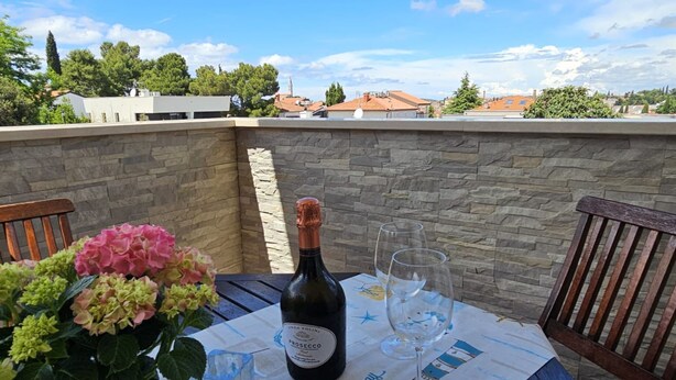 Egy asztal, rajta egy üveg Prosecco, két pohár és egy virágváz egy teraszon, amelynek kőfalai vannak, és ahonnan kilátás nyílik az épületekre.