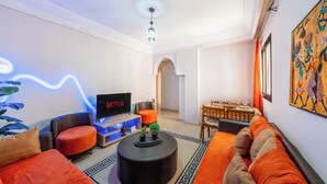 A modern living room with a flat-screen TV, orange seating, a coffee table, and a dining area with a table and chairs.