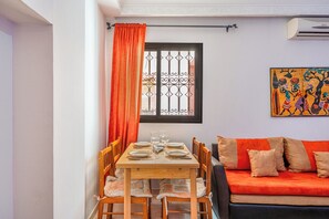 Un coin repas avec une table en bois dressée pour quatre personnes, un canapé avec des coussins orange et une fenêtre ornée de barreaux en fer décoratifs.