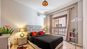 A bedroom with a bed, bedside lamps, a window with curtains, a hanging rack, and a potted plant.