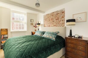 A bedroom with a green quilted bedspread, a wooden dresser, a brick wall, and a window with blinds. - Fitzrovia Flat in 1760s House Central London (London)