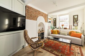 En stue med murstensvæg, hvide skabe, en sofa med orange puder, en rød gulvtæppe med mønster, og et fjernsyn på et skab.