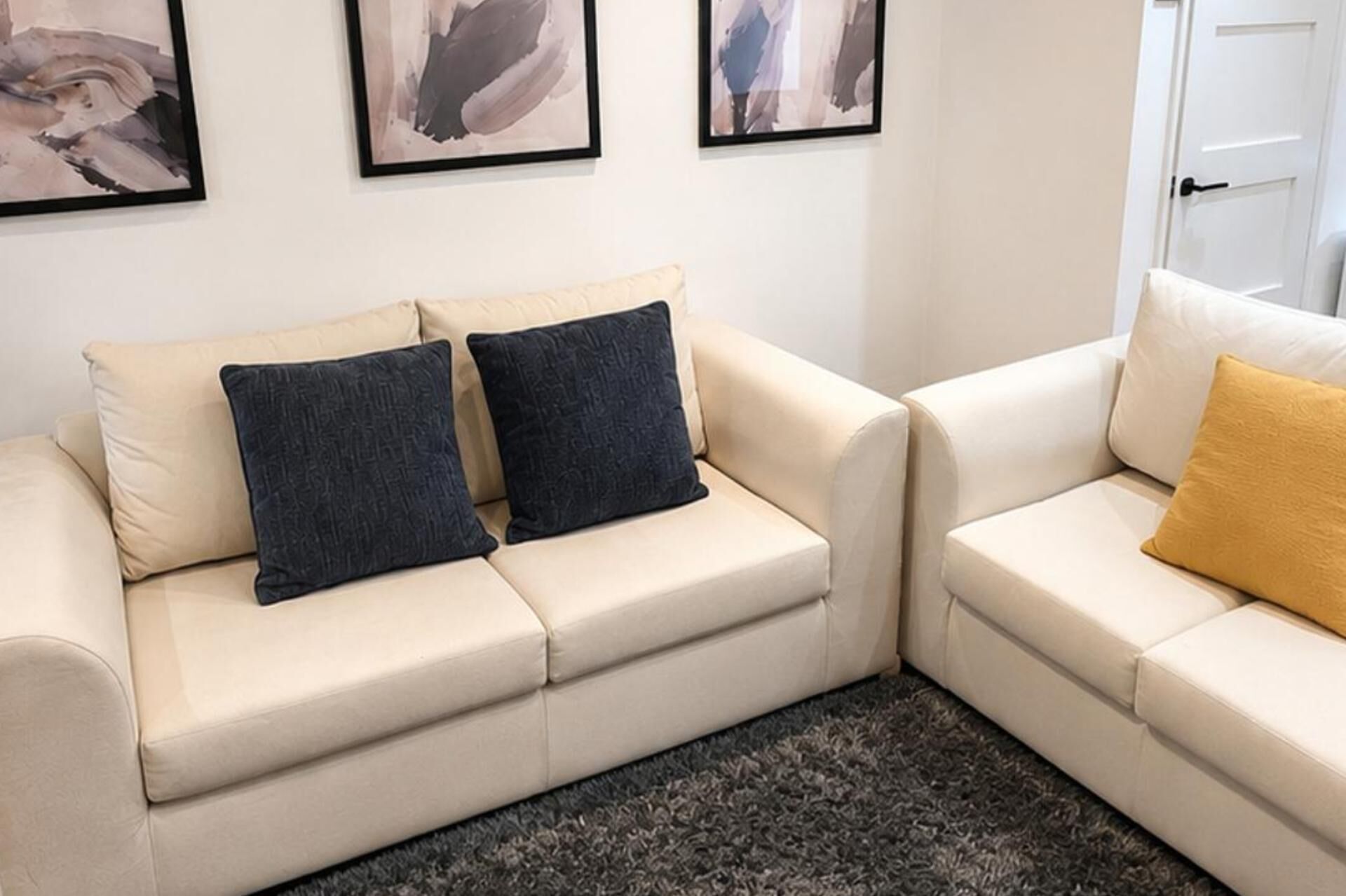 Un divano beige con due cuscini scuri e un cuscino giallo, un tappeto scuro e quadri astratti alle pareti.