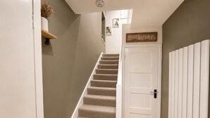 A staircase leading to a door with a 'Welcome Home' sign, a small shelf with dried flowers, and a wall-mounted picture frame. - Chester Charm - Spacious 3 Bedroom House (Chester)