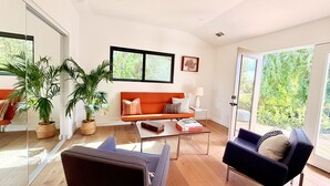 Una sala de estar moderna con sofá, mesa de centro, sillas, ventanas grandes y plantas en macetas.