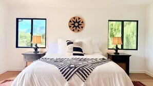A bedroom with a bed, two nightstands, a wall clock, and two windows with views of greenery. - Ojai Valley Luxury Hillside Estate (Ojai)