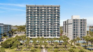 Un hôtel de grande hauteur, avec de nombreux étages, un espace piscine doté de chaises longues et un ciel dégagé.