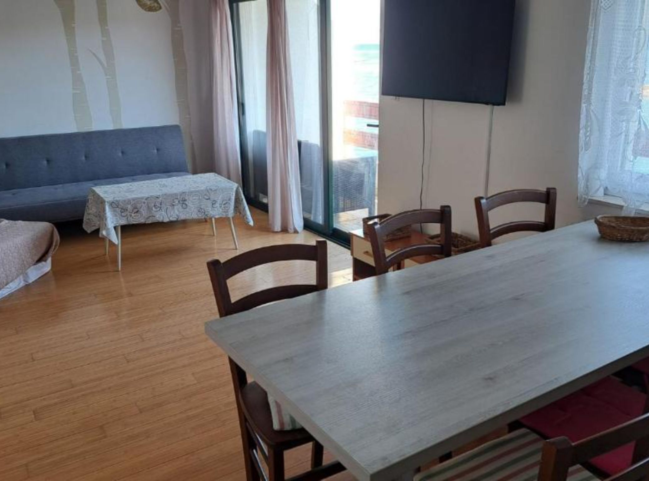 Un comedor con una mesa y sillas, un sofá, una mesita con mantel estampado y un televisor grande montado en la pared.