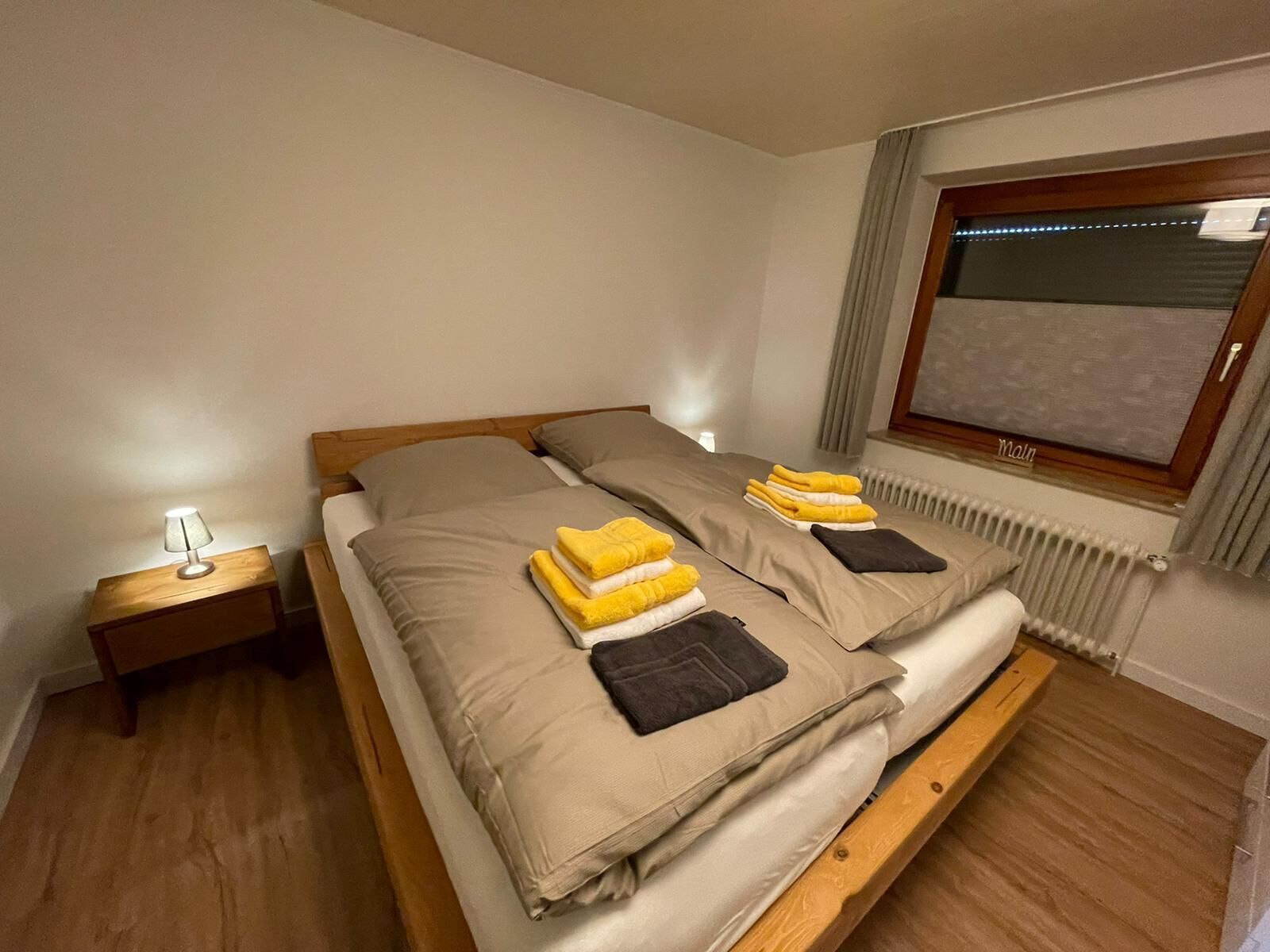 Ein Doppelbett mit beige farbenem Bettzeug und zwei Kissen, ein Nachttisch mit Lampe und ein Fenster mit Vorhängen.