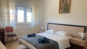 Ein Schlafzimmer mit einem Bett, einem Sessel, einem Nachttisch, einer Lampe und einem Fenster mit Vorhängen.