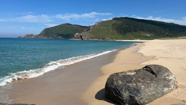 Ein Strand mit einem großen Felsen im Vordergrund, einem sandigen Ufer und einer Hügelkette mit Bäumen im Hintergrund.