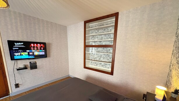A room with a bed, a television mounted on the wall displaying a streaming service, a window with a decorative frame, and a small table with a lamp. - ✨Quiet & cosy apartment - Just like home (Raon-l'Étape)