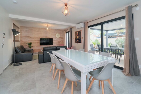 Un salon moderne avec une table à manger en verre, des chaises grises, un canapé, un téléviseur et une grande fenêtre avec des rideaux.