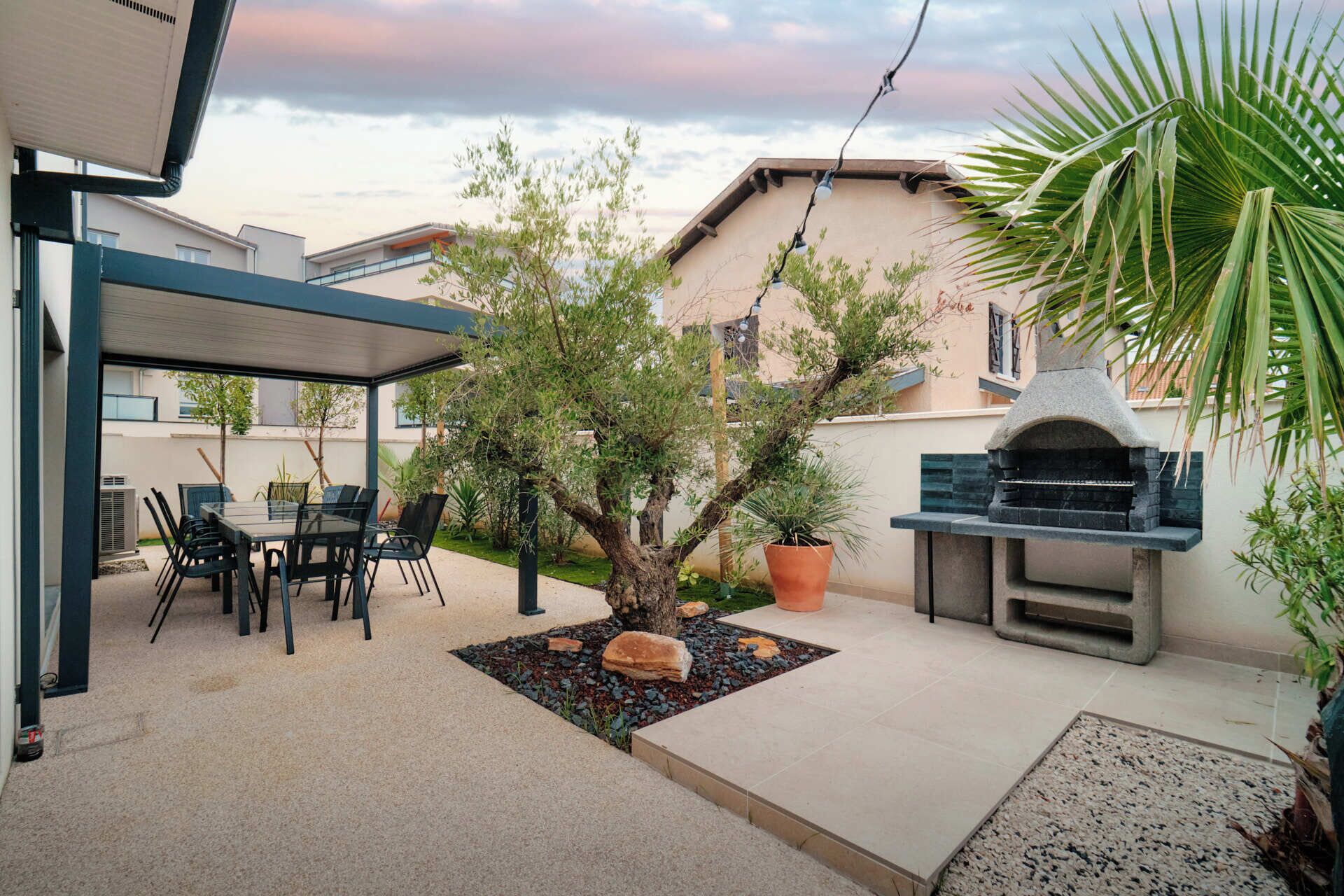 Une terrasse couverte avec une table et des chaises, un petit arbre, un barbecue et une plante en pot.