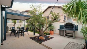 Une terrasse couverte avec une table et des chaises, un petit arbre, un barbecue et une plante en pot.