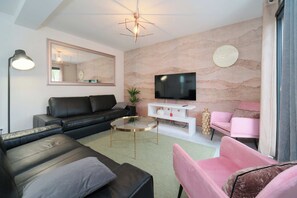 A modern living room with a black leather sofa, a pink armchair, a round glass coffee table, a white entertainment unit, and a flat-screen TV mounted on the wall.