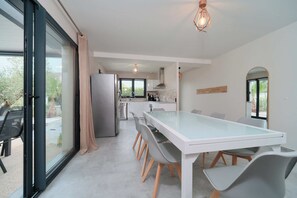 Une cuisine moderne dotée d’un grand îlot central, d’un coin repas avec une table en verre et d’une porte-fenêtre coulissante donnant sur une terrasse.