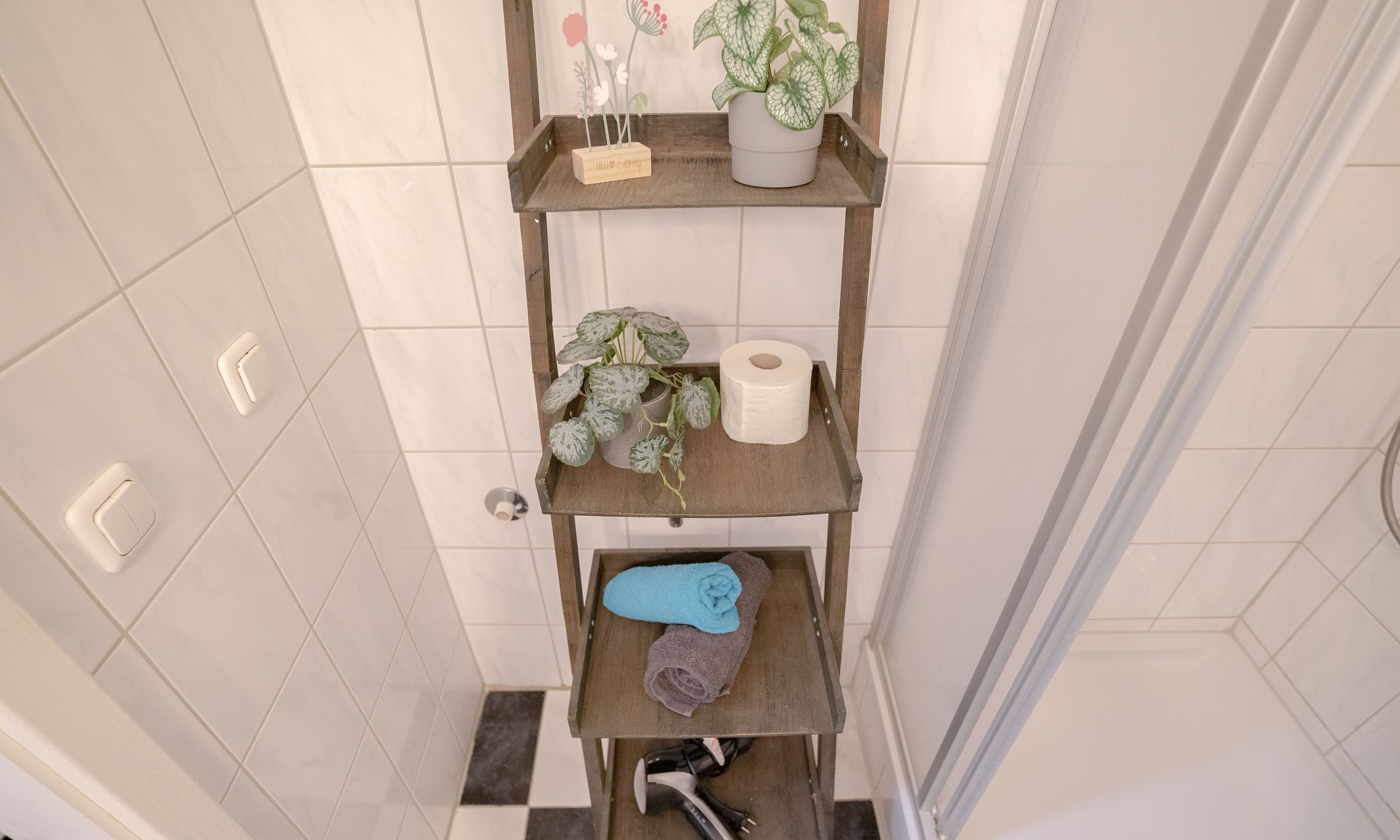 Kylpyhuone, jossa on wc, pyyheteline ja hylly, jossa on hygieniatuotteita ja kasvi.