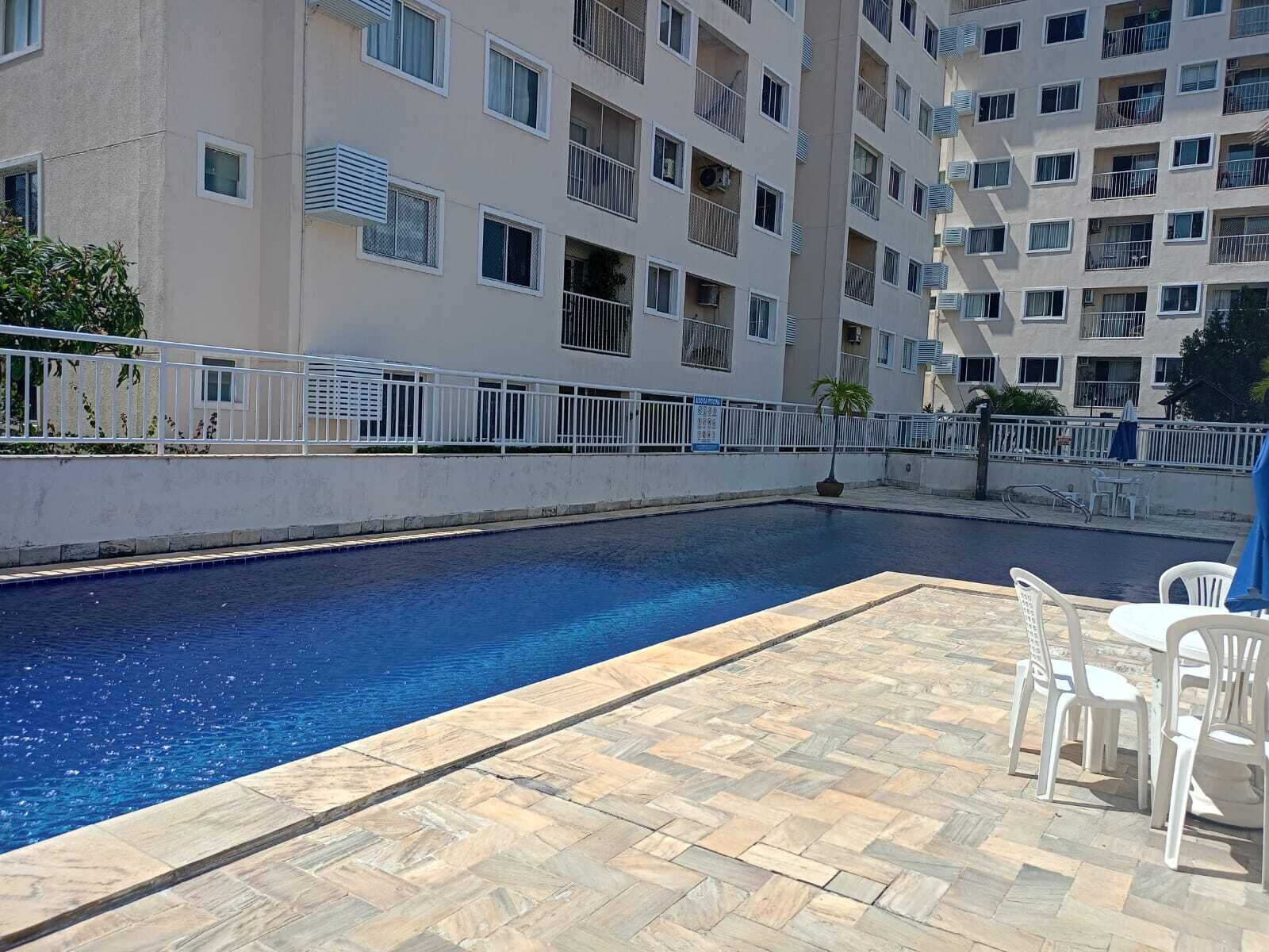A swimming pool with a tiled deck and white chairs, surrounded by a high-rise building with balconies.