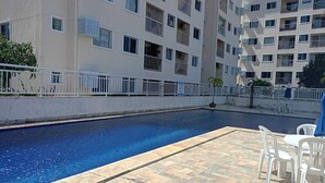 Una piscina amb terrassa de rajola i cadires blanques, envoltada d'un edifici de pisos amb balcons.
