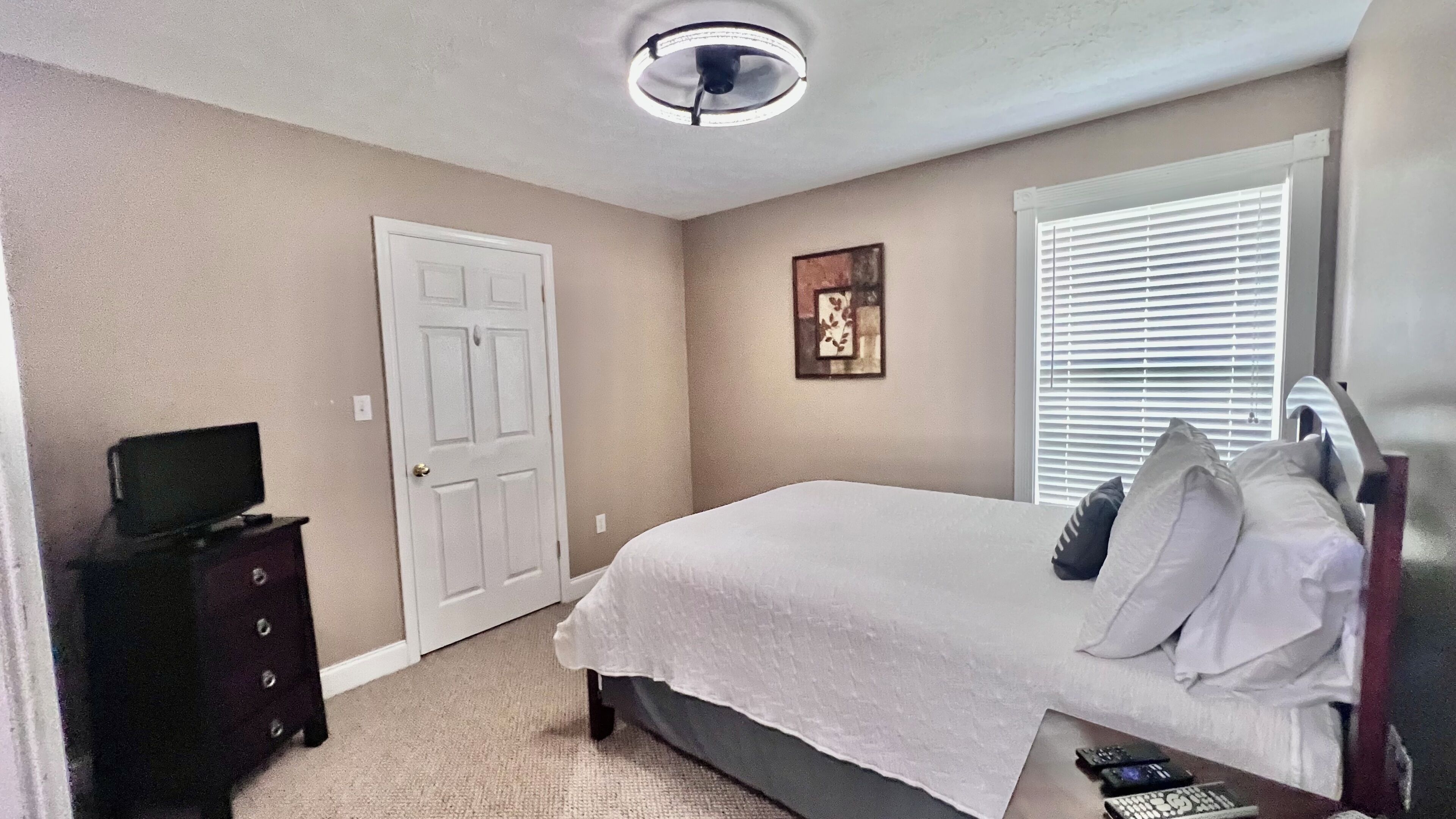 A bedroom with a bed, a dresser, a television, a ceiling fan, a window with blinds, and a picture on the wall.