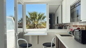 A modern kitchen with a washing machine, a microwave, and a window with a view of palm trees. - Costa Adeje Sunset Ocean View Garden Studio B (Adeje)