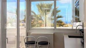 A modern kitchen with a view of palm trees and the sea. There are two white bar stools and a white countertop with a sink. - Costa Adeje Sunset Ocean View Garden Studio B (Adeje)