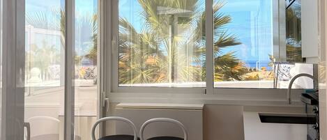 Une cuisine moderne avec vue sur les palmiers et la mer. On y trouve deux tabourets de bar blancs et un plan de travail blanc avec un évier.