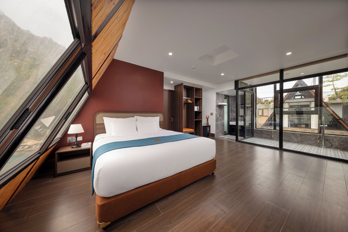 Une chambre d’hôtel moderne avec un grand lit, une table de chevet en bois, un bureau et une vue sur les montagnes par une grande fenêtre.