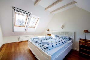 Une chambre avec un plafond en pente, deux fenêtres, un lit avec une couette à pois, une table de chevet avec une lampe et un radiateur.