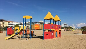 Ein Spielplatz am Strand mit Rutschen und Klettergerüsten, vor einer Kulisse aus Gebäuden und blauem Himmel.