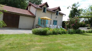 A two-story house with a red-tiled roof, a garden with various plants and flowers, and a patio with white chairs and a yellow umbrella.