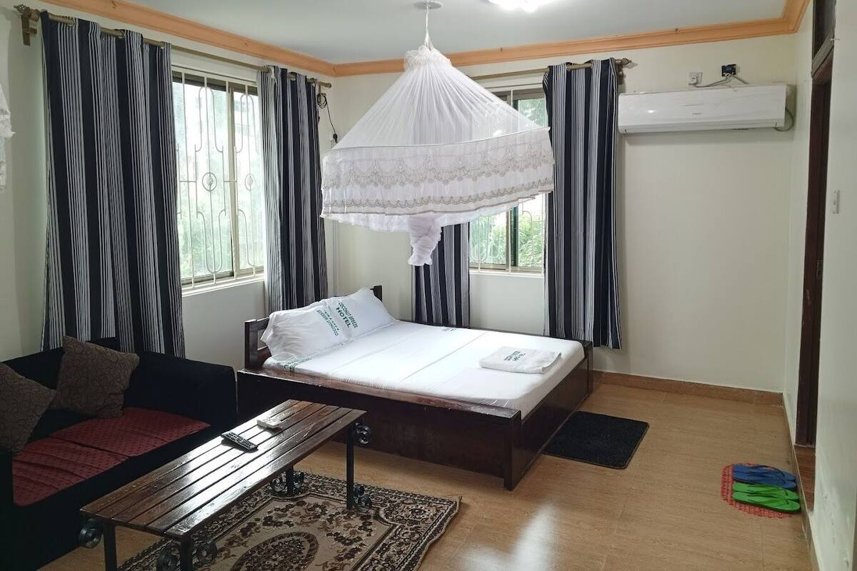 Un dormitorio con cama, mesita de noche, mosquitero colgado y ventana con cortinas.