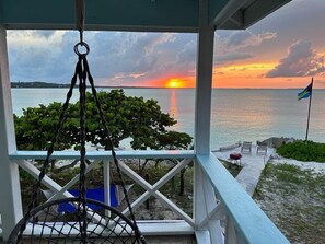 A view from a porch overlooking a sunset over a body of water, with a flag and outdoor seating visible.