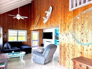 A spacious living room with a ceiling fan, a grey armchair, a wooden wall, a television, and a bookshelf.