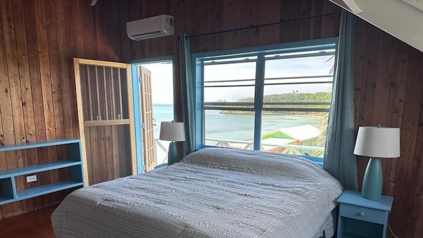 A bedroom with a large bed, wooden walls, a view of the sea, and a window with a view of buildings. - Cottage style living directly on the Ocean (Georgetown)
